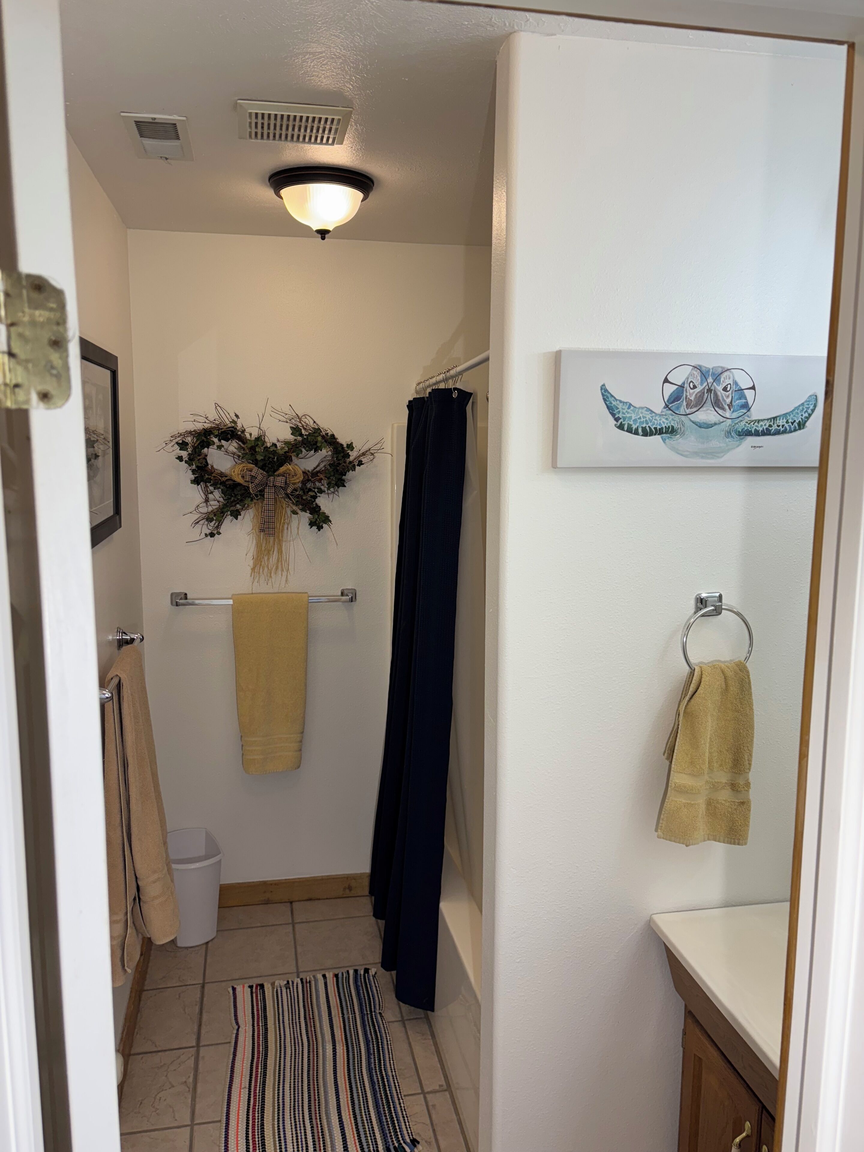 A bathroom with a shower curtain, a towel rack, a framed artwork, and a small trash bin.