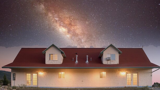 A two-story house with a red roof, two small attached structures on the roof, and a well-lit front entrance. - Hacienda Roja - The Juniper #5 at Wind Walker Homestead (Spring City)