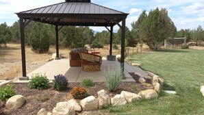 A gazebo with a concrete floor and a metal roof, surrounded by a landscaped garden with rocks and flowers. - Hacienda Roja - The Juniper #5 at Wind Walker Homestead (Spring City)