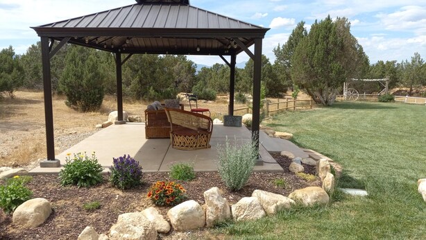 A gazebo with a concrete floor and a metal roof, surrounded by a landscaped garden with rocks and flowers. - Hacienda Roja - The Sage #6 at Wind Walker Homestead (Spring City)