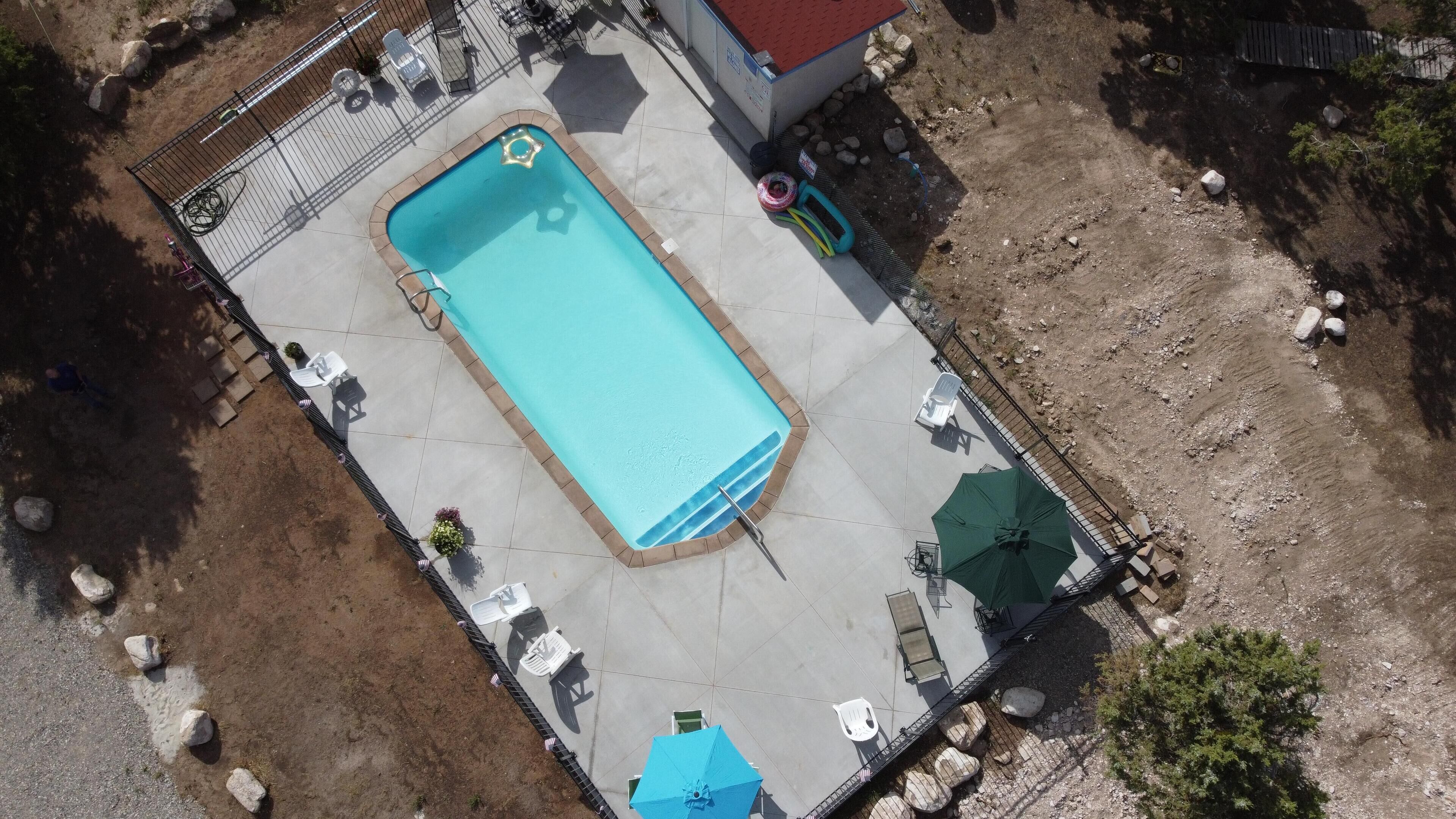 Vista aérea de una piscina con solárium empedrado, rodeada de mobiliario exterior y una zona vallada.