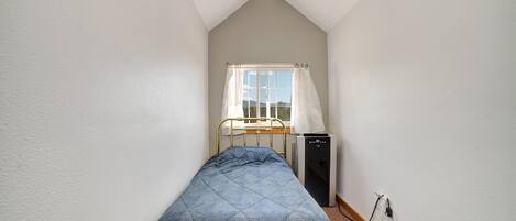 A small, narrow room with a single bed, a window with curtains, and a small heater or appliance against the wall.