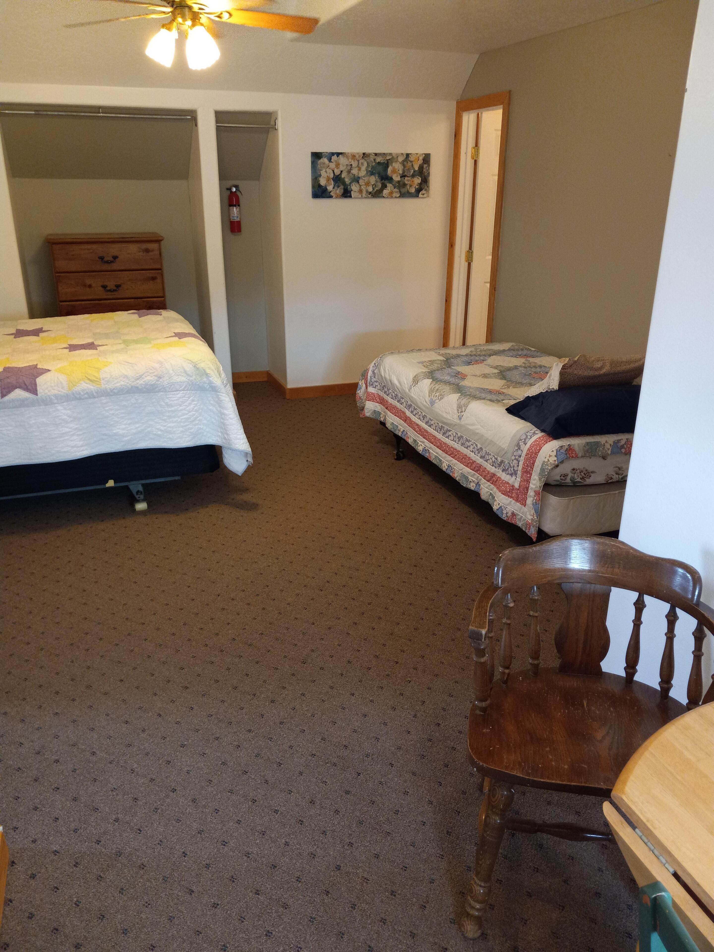 A bedroom with two beds, a wooden chair, a ceiling fan, and a painting on the wall.