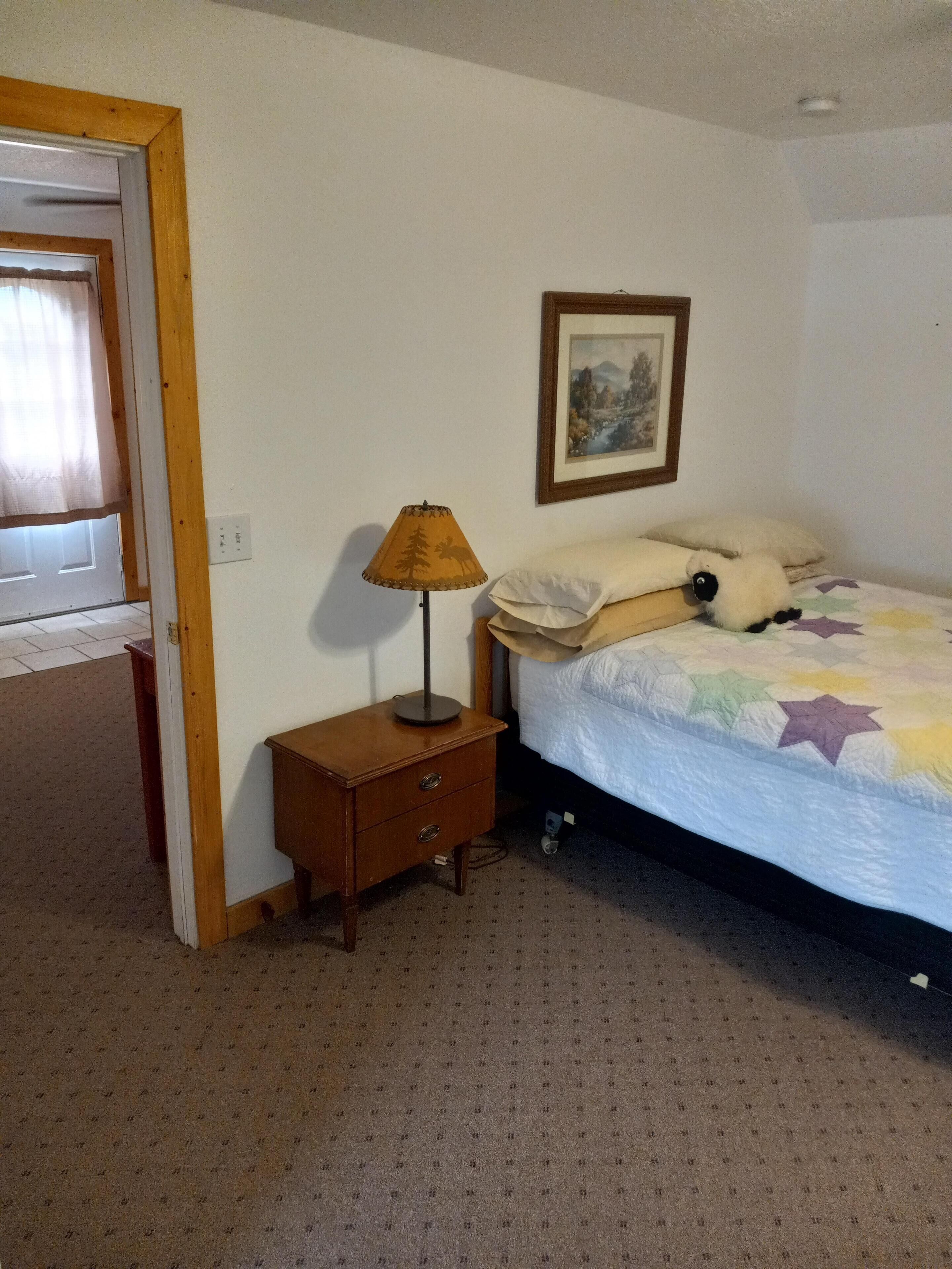 Une chambre à coucher comprenant un lit, une table de chevet, une lampe, une porte et un tableau accroché au mur.