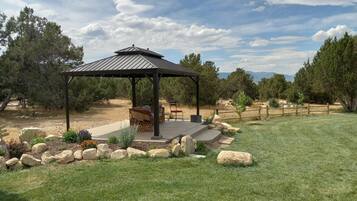 A gazebo with a metal roof, surrounded by a stone border, in a grassy area with trees and a wooden fence in the background.