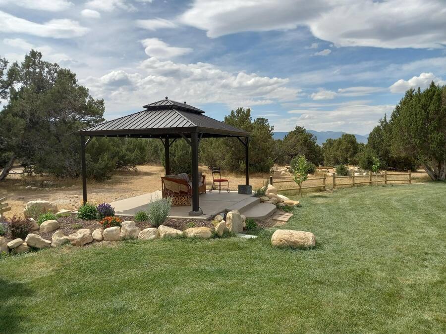 A gazebo with a metal roof, surrounded by a stone border, in a grassy area with trees and a wooden fence in the background.