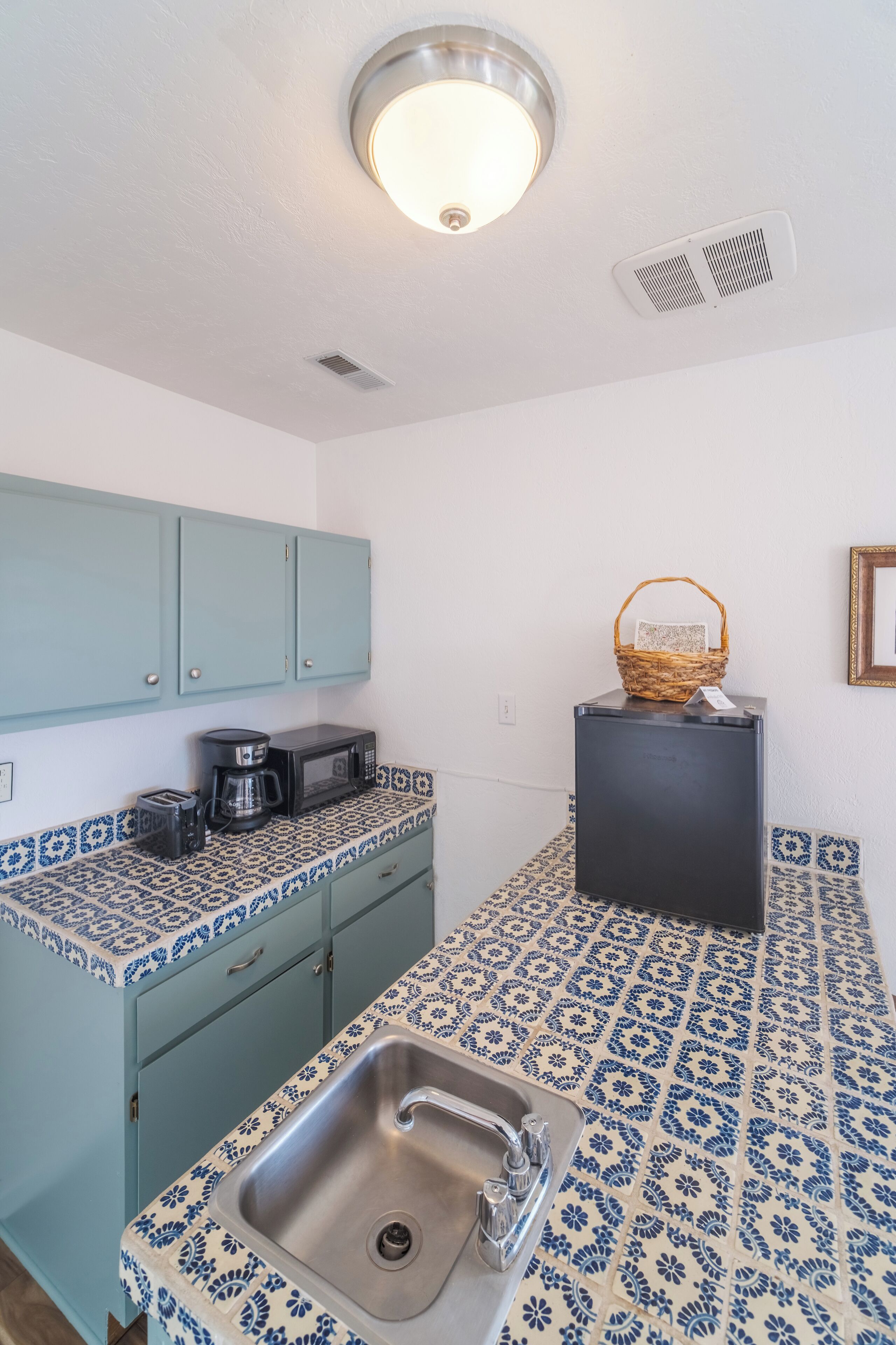Una cocina con un lavabo, microondas y cafetera sobre la encimera. Hay una cesta en una pequeña mesa y un cuadro en la pared.