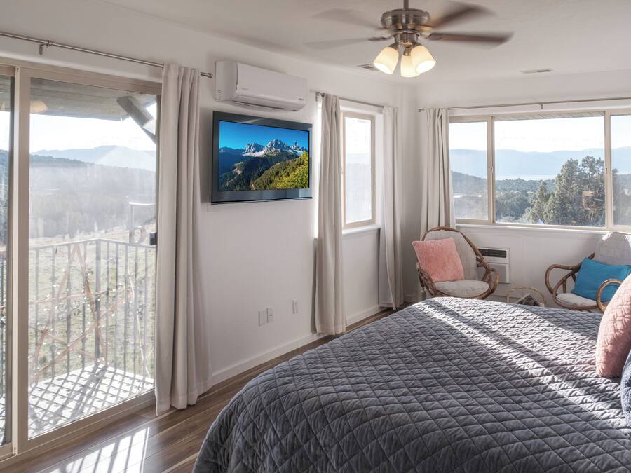 Un dormitorio con una cama grande, un ventilador de techo, un televisor en la pared y un balcón con vistas a la montaña.