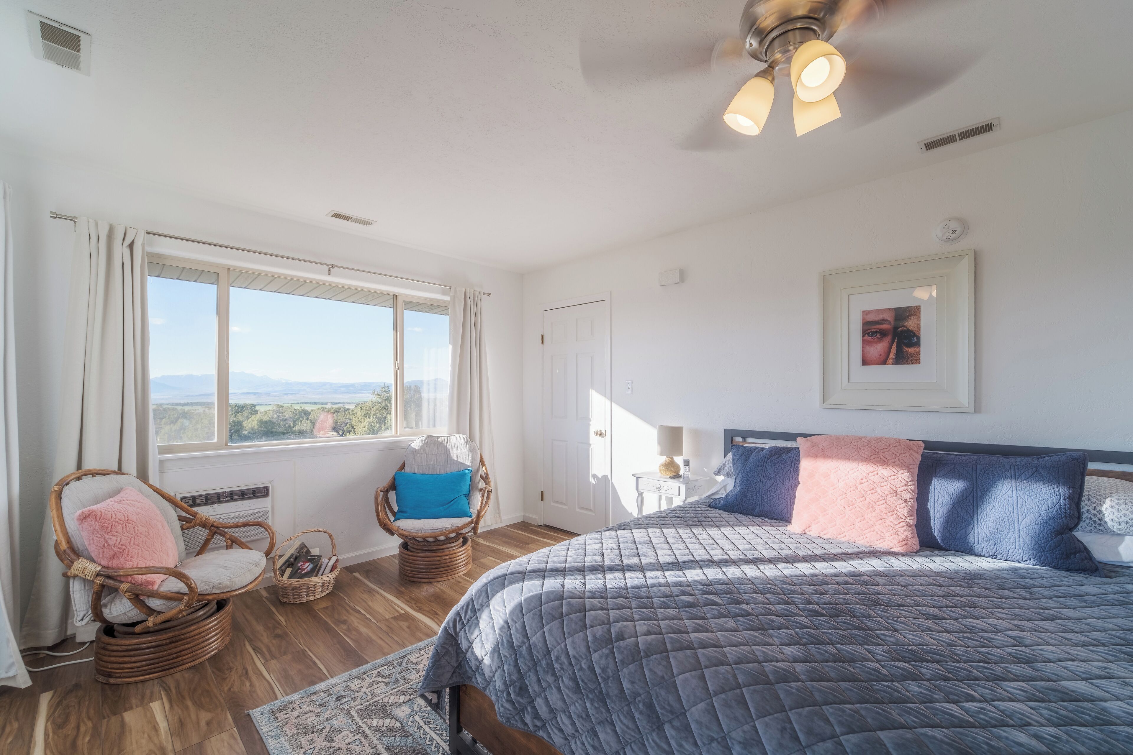 A bedroom with a large bed, a wicker chair, a small table, and a view of the outdoors.