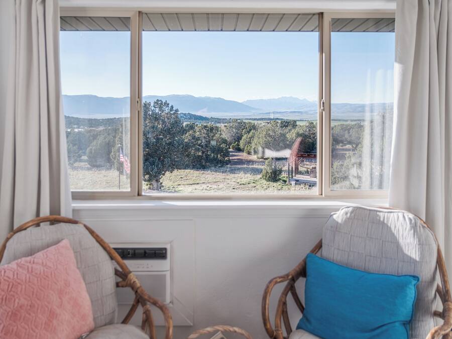 A room with a large window overlooking a landscape with mountains and trees. There are two wicker chairs with cushions.