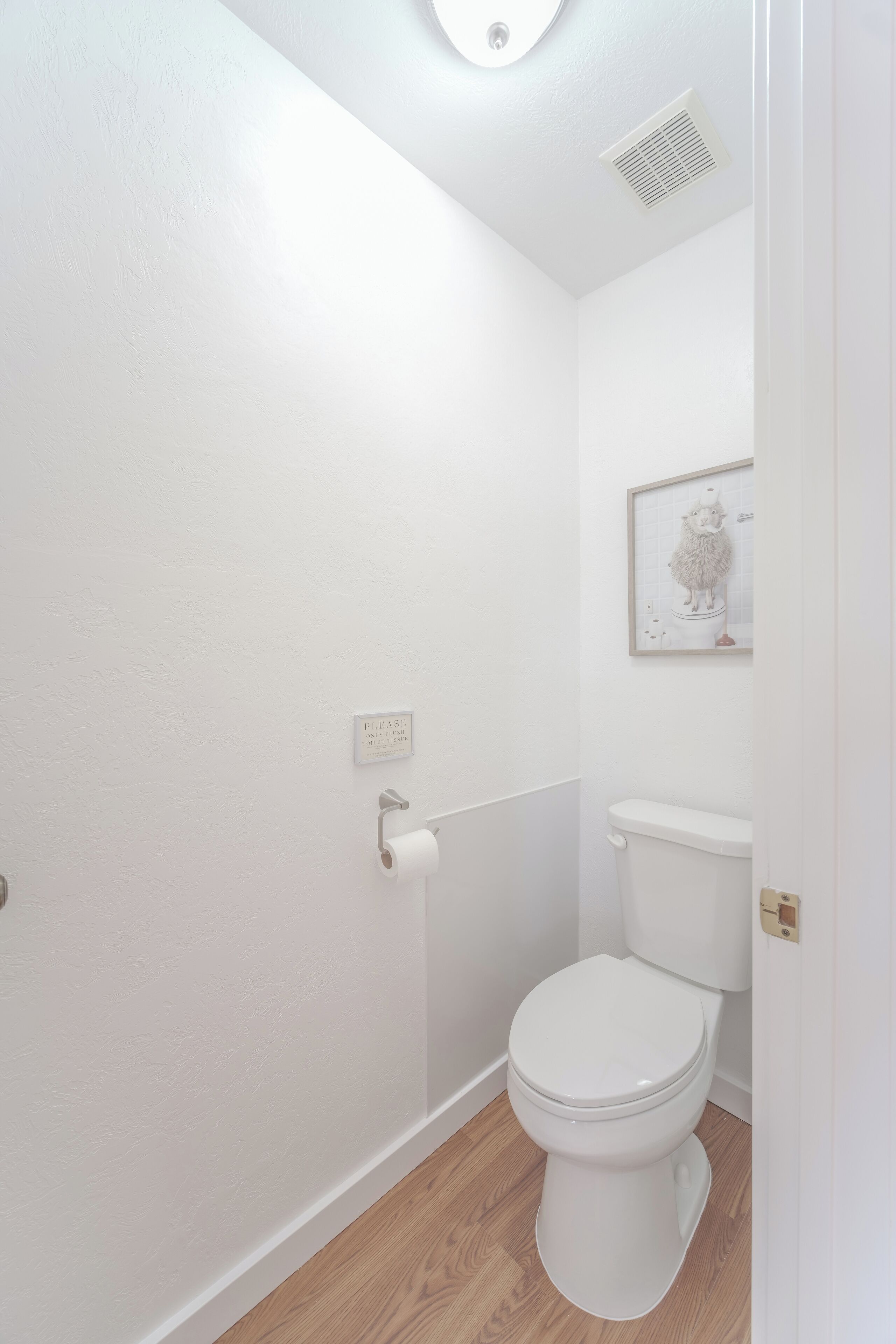 A small, white bathroom with a toilet, a toilet paper holder, and a framed picture on the wall.