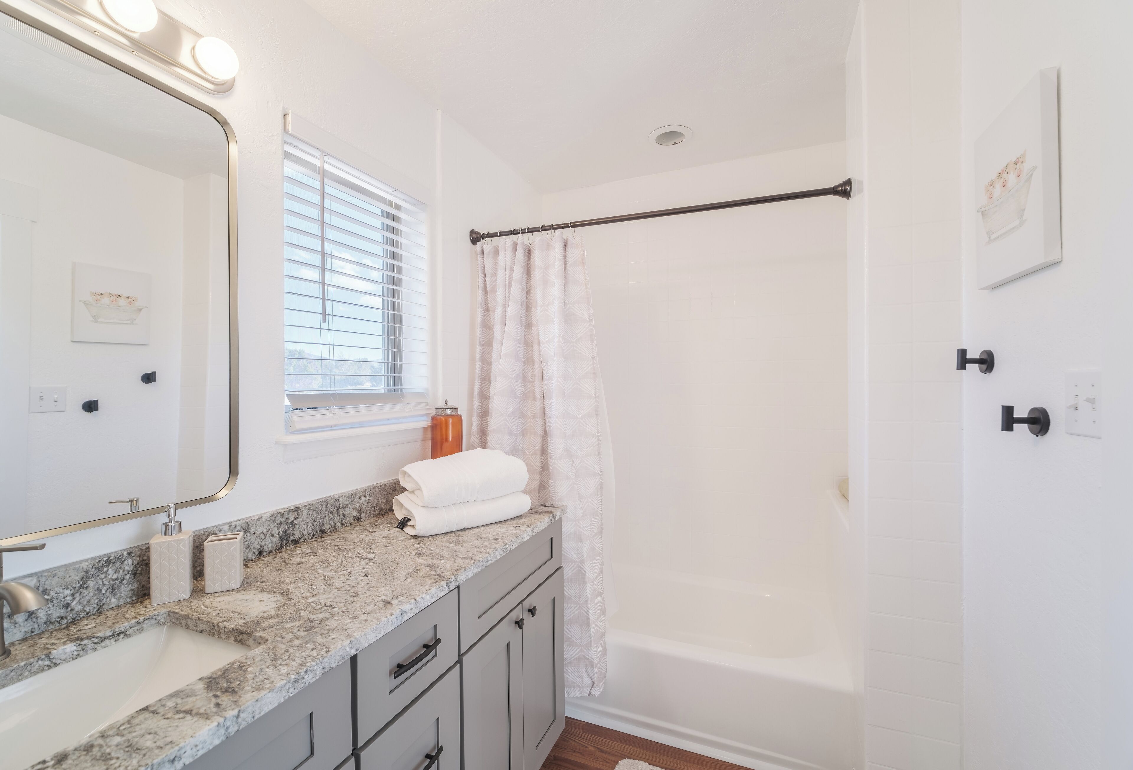 A bathroom with a granite countertop, a large mirror, a window with blinds, a shower curtain, and a bathtub.