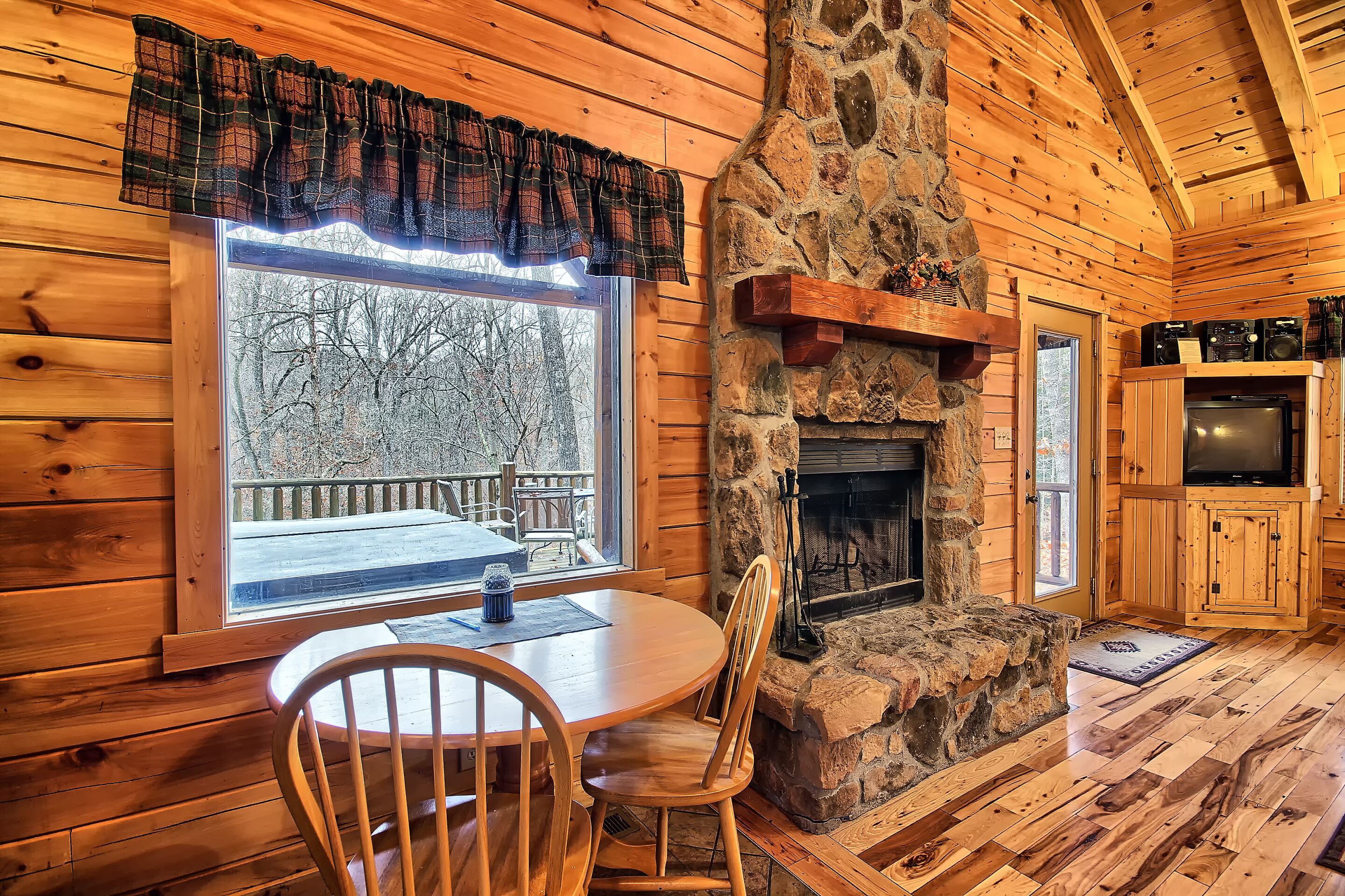 A cozy cabin interior with a stone fireplace, wooden walls, a dining table with chairs, and a large window overlooking a snowy deck.