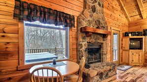 A cozy cabin interior with a stone fireplace, wooden walls, a dining table with chairs, and a large window overlooking a snowy deck. - Grant's Escape - Couples Cabin in Hocking Hills (Logan)