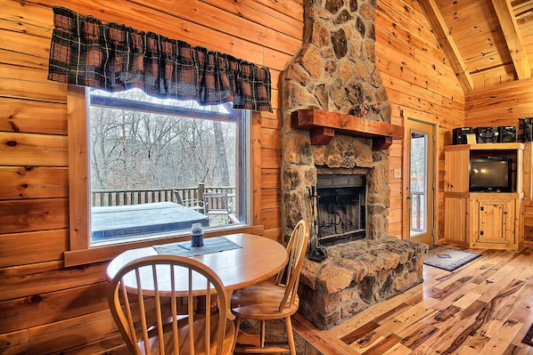 A cozy cabin interior with a stone fireplace, wooden walls, a dining table with chairs, and a large window overlooking a snowy deck.