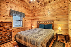 A wooden cabin room with a bed, dresser, window with blinds, and two bedside lamps.