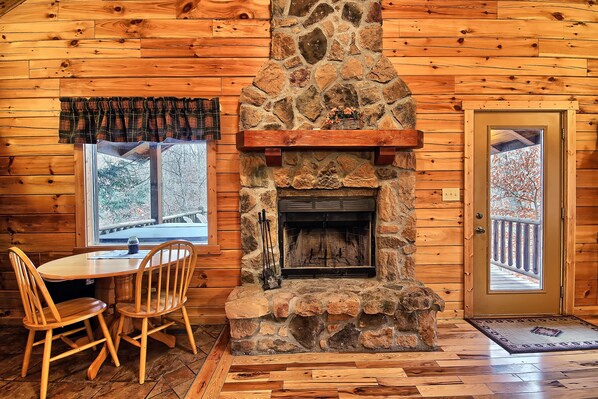 A stone fireplace with a mantel, wooden walls, a dining table with chairs, and a door with a view of a balcony.