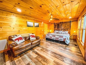 Interior de una cabaña de madera con sofá de cuero, una cama con manta de cuadros y paredes de madera.