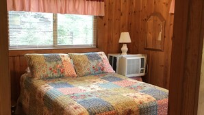 A bedroom with a wooden paneled wall, a bed with a floral quilt, a ceiling fan, a window with blinds, and a white air conditioning unit. - Fence Lake Retreat w/ dock (Lac du Flambeau)