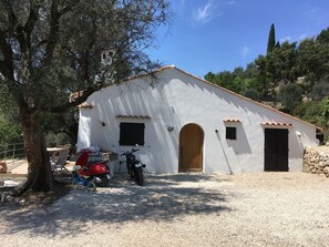 A white building with a tiled roof, a wooden door, and a small window. There is a tree to the left and a motorcycle parked in front.