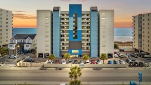 Ein mehrstöckiges Hotel mit einem zentralen blauen Abschnitt, Parkplatz und Blick auf den Strand.
