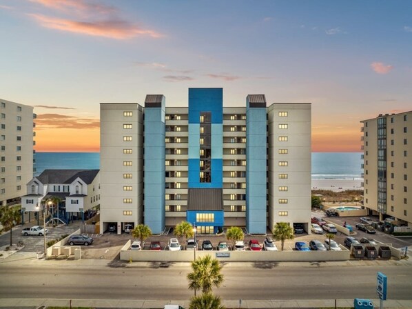 Hotel com vários andares, uma seção central azul, estacionamento e vista para a praia.