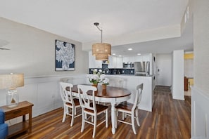 Uma cozinha moderna com armários brancos, uma área de refeições com mesa de madeira e cadeiras brancas, e um lustre pendente.