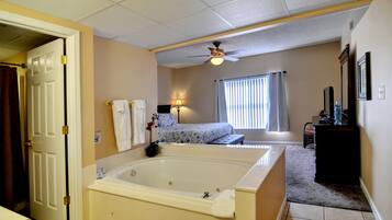 A bedroom with a large bathtub, a bed with a floral bedspread, a ceiling fan, and a window with curtains.