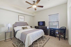 A bedroom with a bed, a desk, a chair, a ceiling fan, a window with blinds, and a wall-mounted TV.