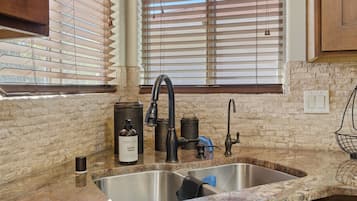 A kitchen with a granite countertop, stainless steel sink, and a window with blinds.