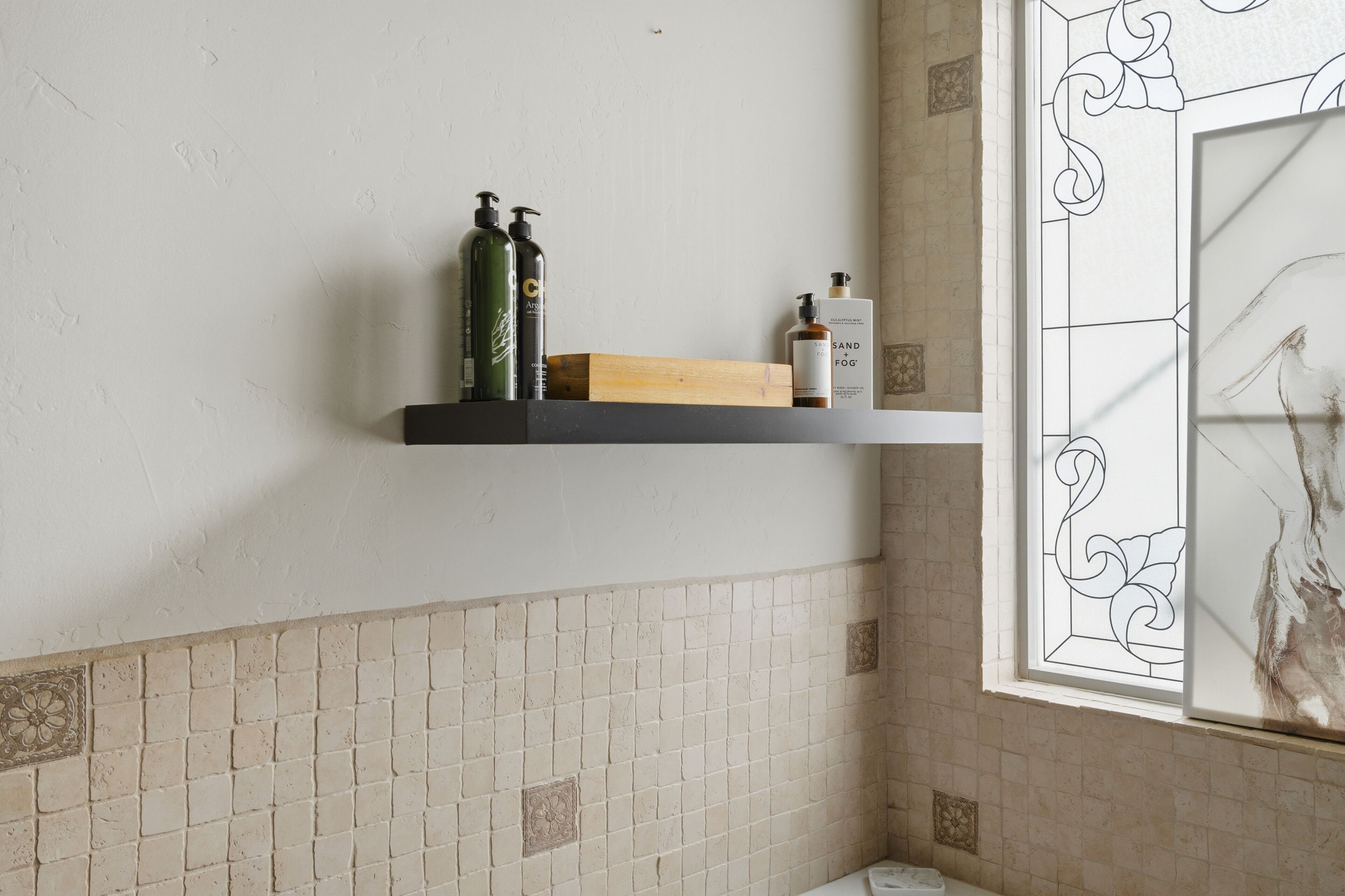 A bathroom with a tiled wall, a shelf with toiletries, and a window with decorative glass.