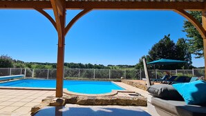 Una piscina exterior techada con pérgola, sillas de descanso y un muro de piedra.