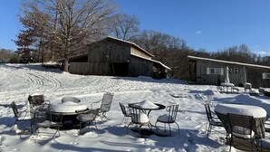 A snow-covered outdoor seating area with metal chairs and tables, a wooden barn, and a small building in the background.