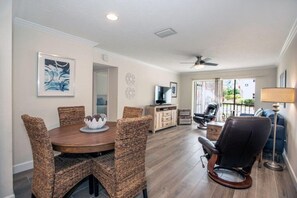 A dining area with a round table and wicker chairs, a massage chair, a TV, and a balcony with a view.