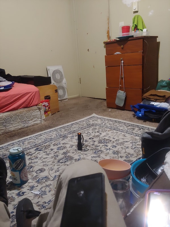 Une pièce encombrée, avec un lit, une commode, un ventilateur et divers objets éparpillés.