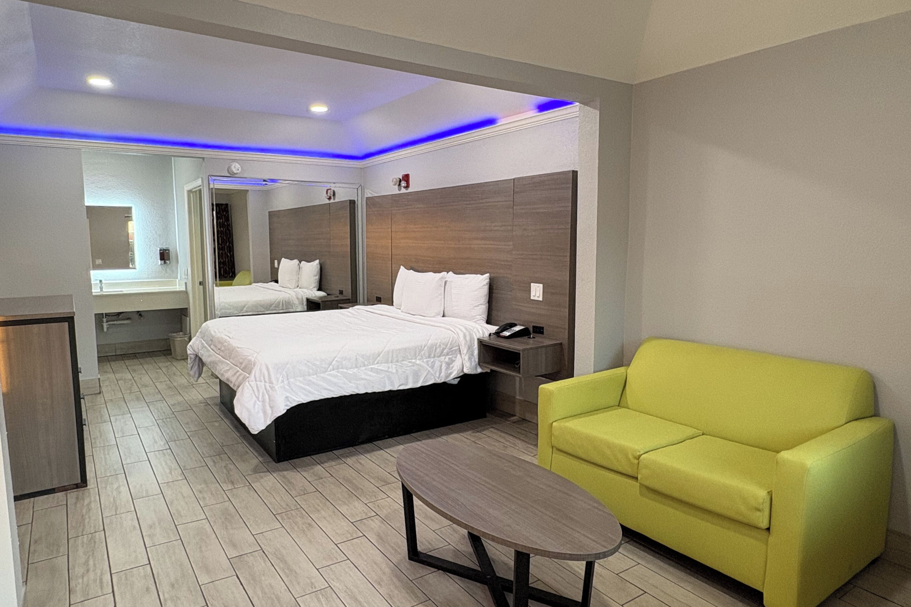 A modern hotel room with a bed, bedside tables, a mirror, a yellow armchair, and a small table.