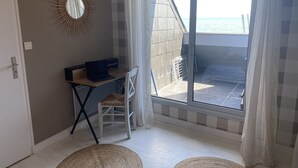 Una habitación con una computadora portátil sobre un escritorio, una silla, un tapete tejido y vista al mar a través de una puerta de vidrio.