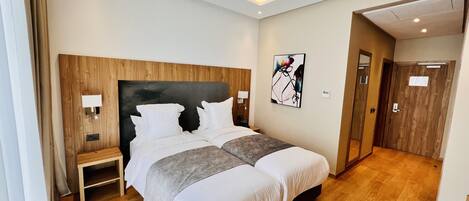Une chambre d’hôtel moderne avec un grand lit, des lampes de chevet, une tête de lit en bois, un tableau au mur et une porte donnant sur une autre pièce.