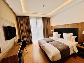 Habitación de hotel con una cama grande, un escritorio, una silla, un televisor y una ventana con cortinas.
