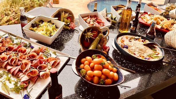 Una mesa de bufé con diversos platos, incluyendo carnes, verduras y salsas, dispuesta al aire libre con sombrillas y una piscina al fondo.