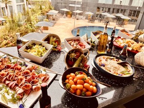 Una mesa de bufé con diversos platos, incluyendo carnes, verduras y salsas, dispuesta al aire libre con sombrillas y una piscina al fondo.