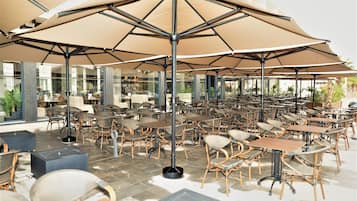 An outdoor café area with multiple tables and chairs, large umbrellas, and a modern architectural design.