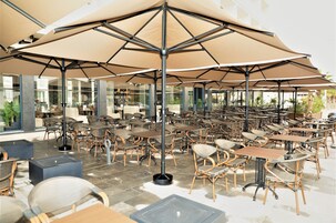 An outdoor café area with multiple tables and chairs, large umbrellas, and a modern architectural design.