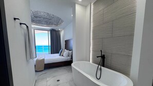 Camera d'albergo moderna con una vasca da bagno ampia, un letto con vista sull'oceano e un balcone con tende.