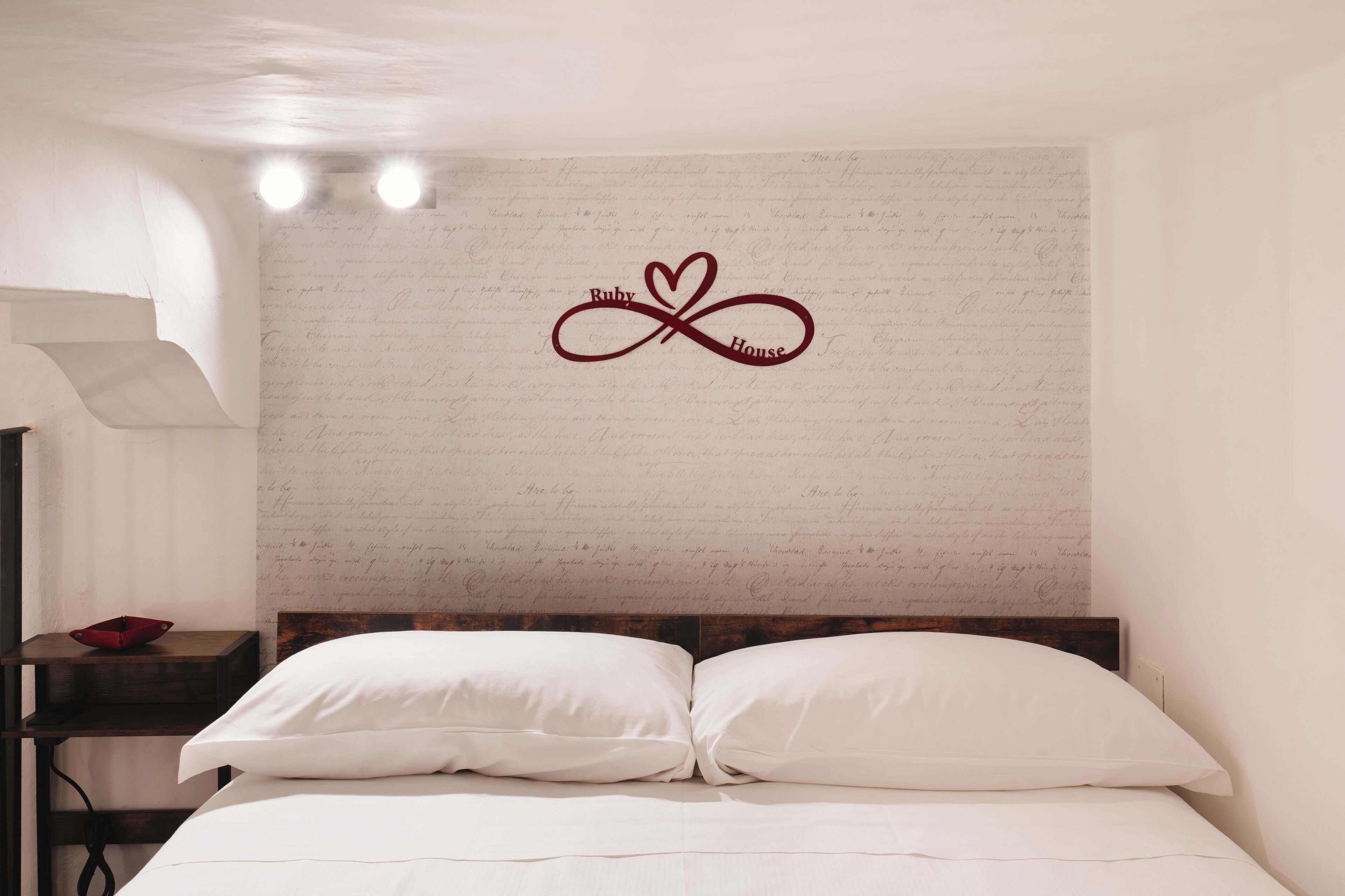 A bedroom with a bed, white pillows, a wooden headboard, a nightstand with a red bowl, and a wall with a heart-shaped infinity symbol.