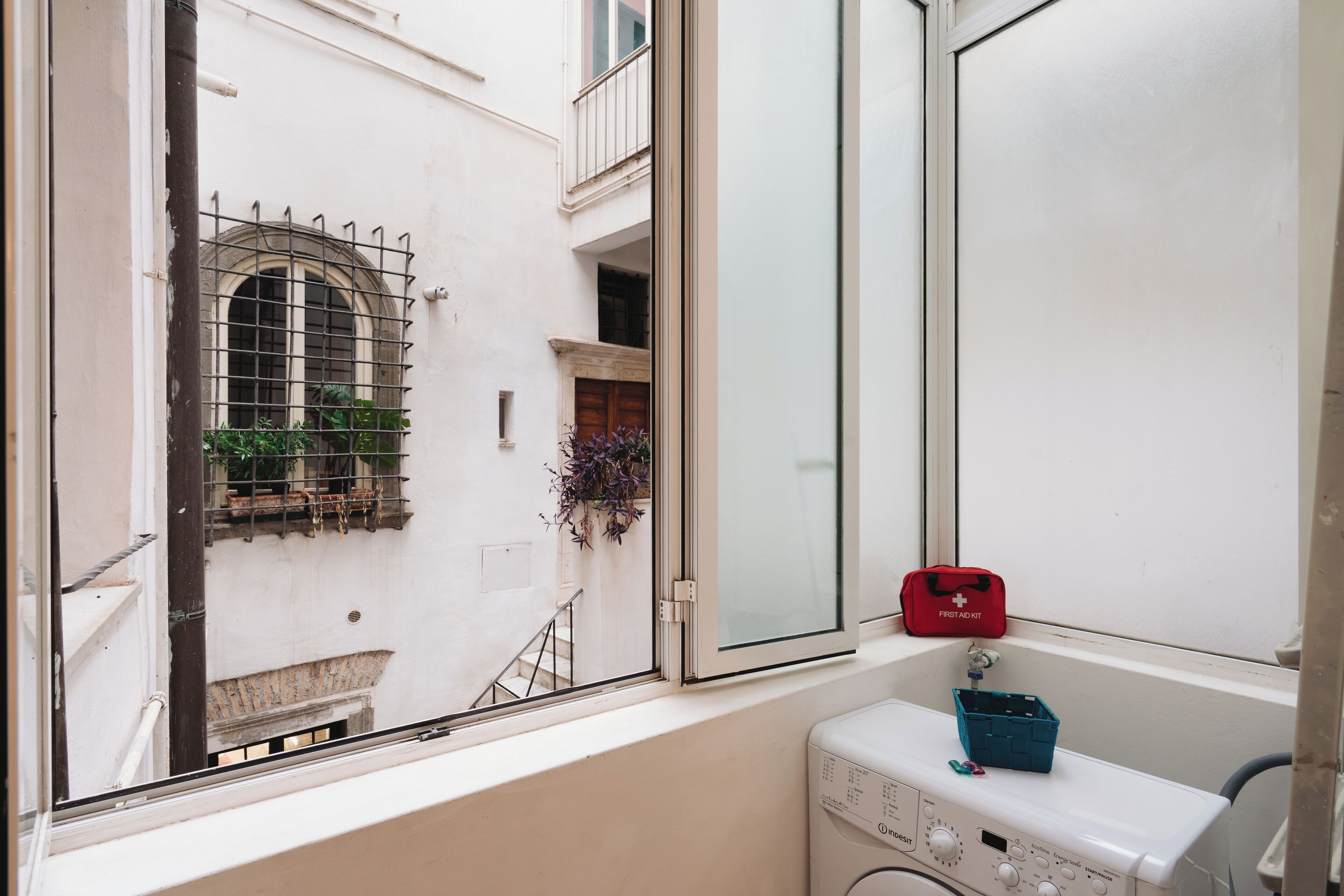 Ein Waschraum mit einer Waschmaschine, einem Fenster mit Blick auf einen Balkon und einer roten Erste-Hilfe-Ausrüstung auf dem Fensterbrett.
