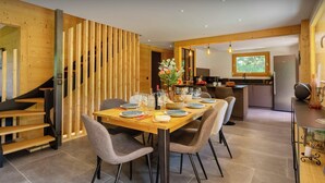 A modern dining area with a wooden table set for six, surrounded by grey upholstered chairs. A kitchen with a built-in oven and a dining area with a wooden partition are visible in the background. - Chalet Hychris - ski holiday between Annecy & the mountains - OVO Network (Alex)