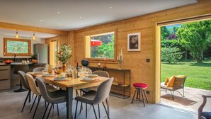 A modern kitchen with wooden walls, a dining area with a table set for four, and a view of a garden through large windows. - Chalet Hychris - ski holiday between Annecy & the mountains - OVO Network (Alex)
