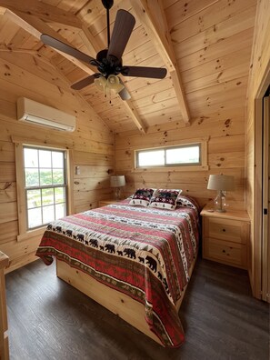 A wooden cabin bedroom with a bed, two bedside tables, a ceiling fan, and two windows.