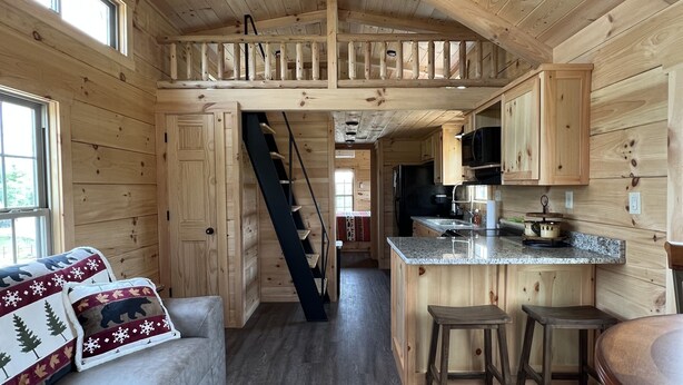 Interior de una cabaña de madera con cocina, comedor y una escalera que sube a un altillo.